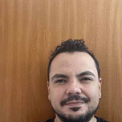 Danilo Valério: homem de cabelo curto escuro e cacheado, barba aparada, brincos pequenos e colar de prata, vestindo camiseta preta, sorrindo levemente em frente a uma parede de madeira clara.